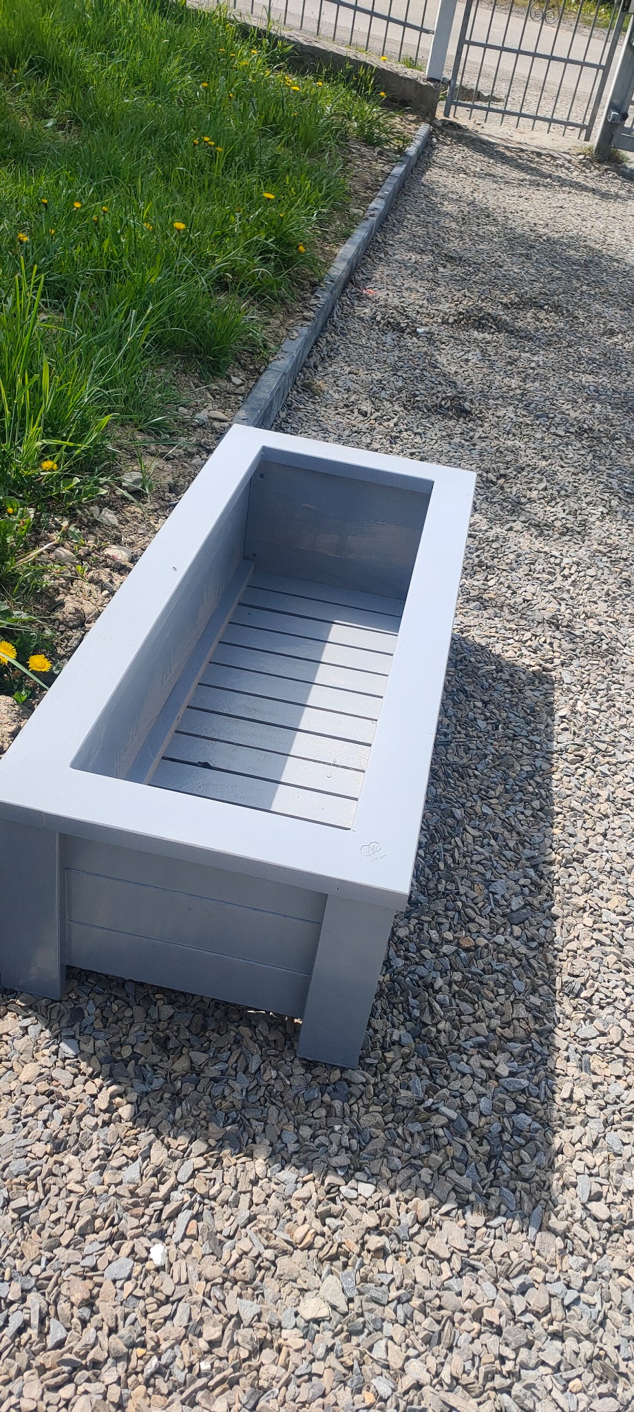 Szara, drewniana skrzynia na kwiaty z lamelowym dnem, ustawiona na żwirowym podłożu, widok z góry.