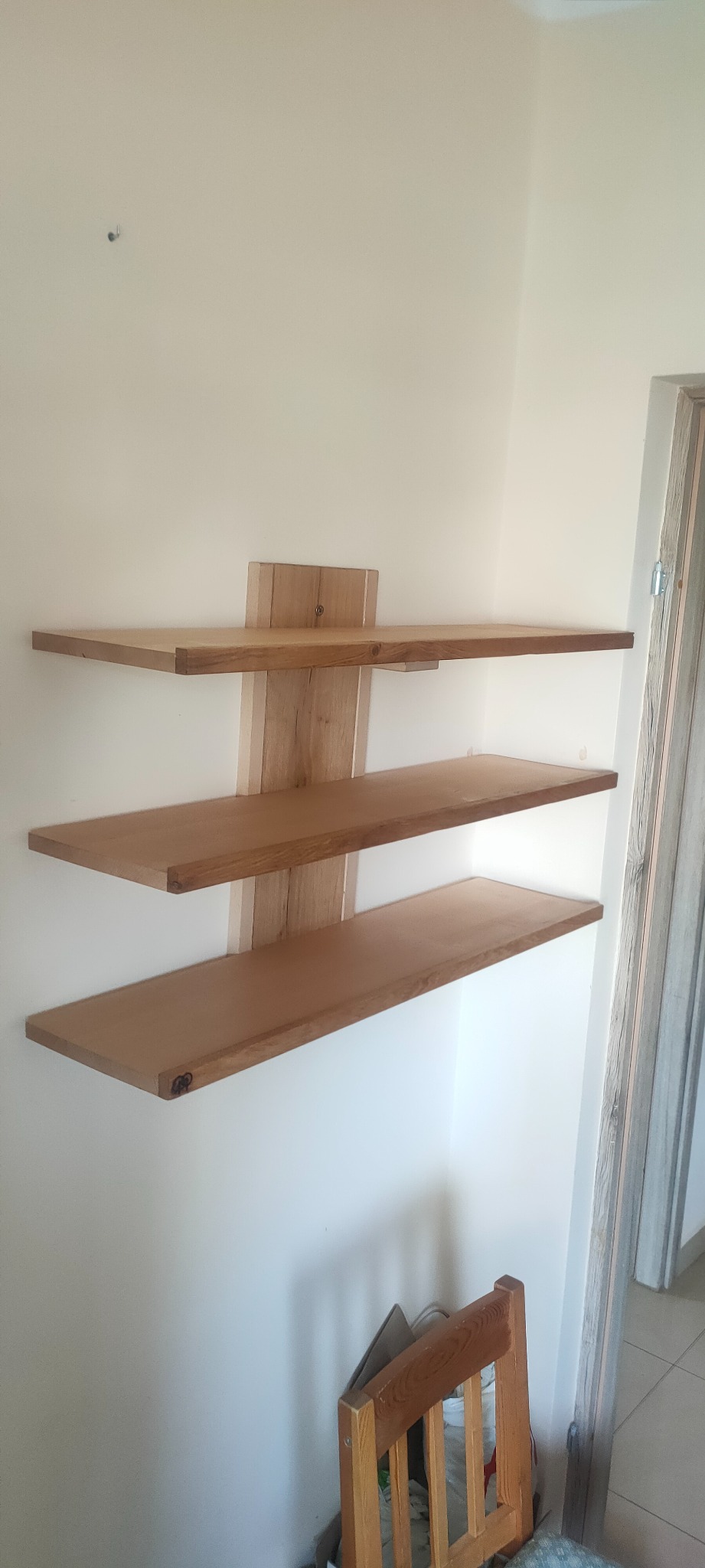 Trzy drewniane półki zamontowane na ścianie, widoczne mocowania, surowe drewno, w tle fragment drzwi i krzesło.