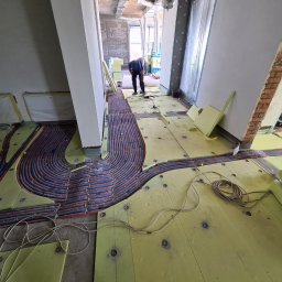 CIEPŁO EKO - Montaż ogrzewania podłogowego: widok z góry na układane rury grzewcze w izolacji z żółtego styropianu, instalator pochylony nad pracą w surowym wnętrzu budynku w trakcie budowy.