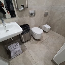 CIEPŁO EKO - Nowoczesna łazienka z wiszącą muszlą klozetową, umywalką z baterią, koszem na śmieci i podestem dla dziecka, wykończona płytkami w odcieniach szarości.