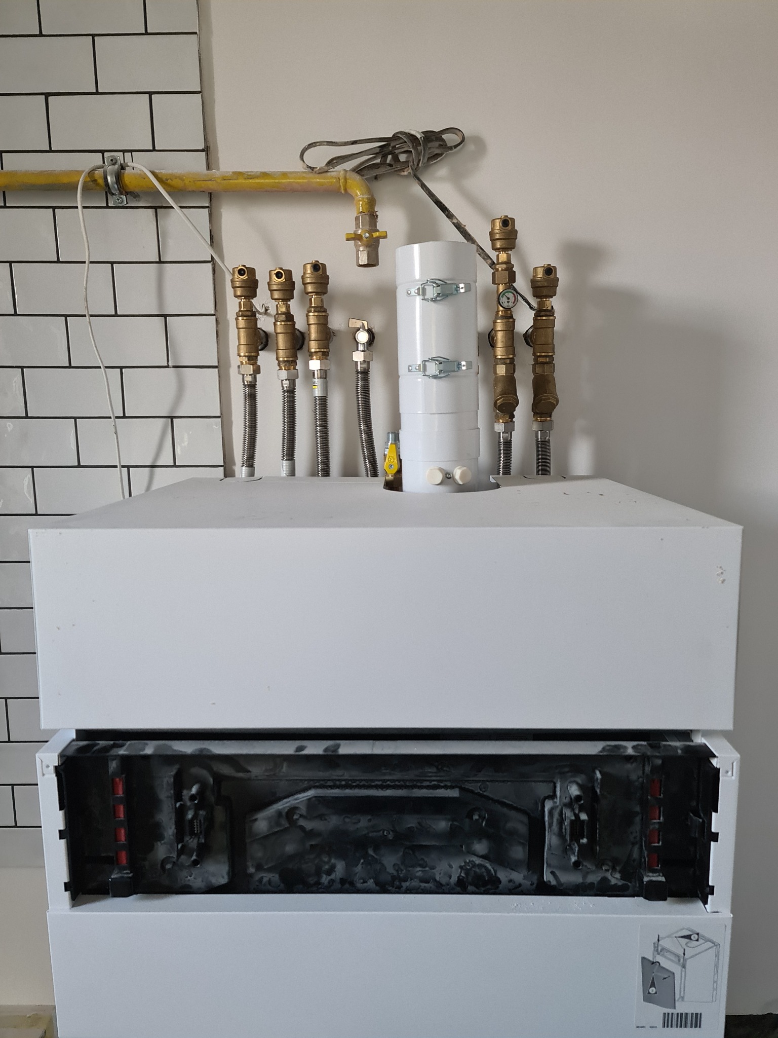 Biały piec grzewczy z widocznymi przyłączami miedzianymi na tle ściany z białej cegły i gładkiej ściany.