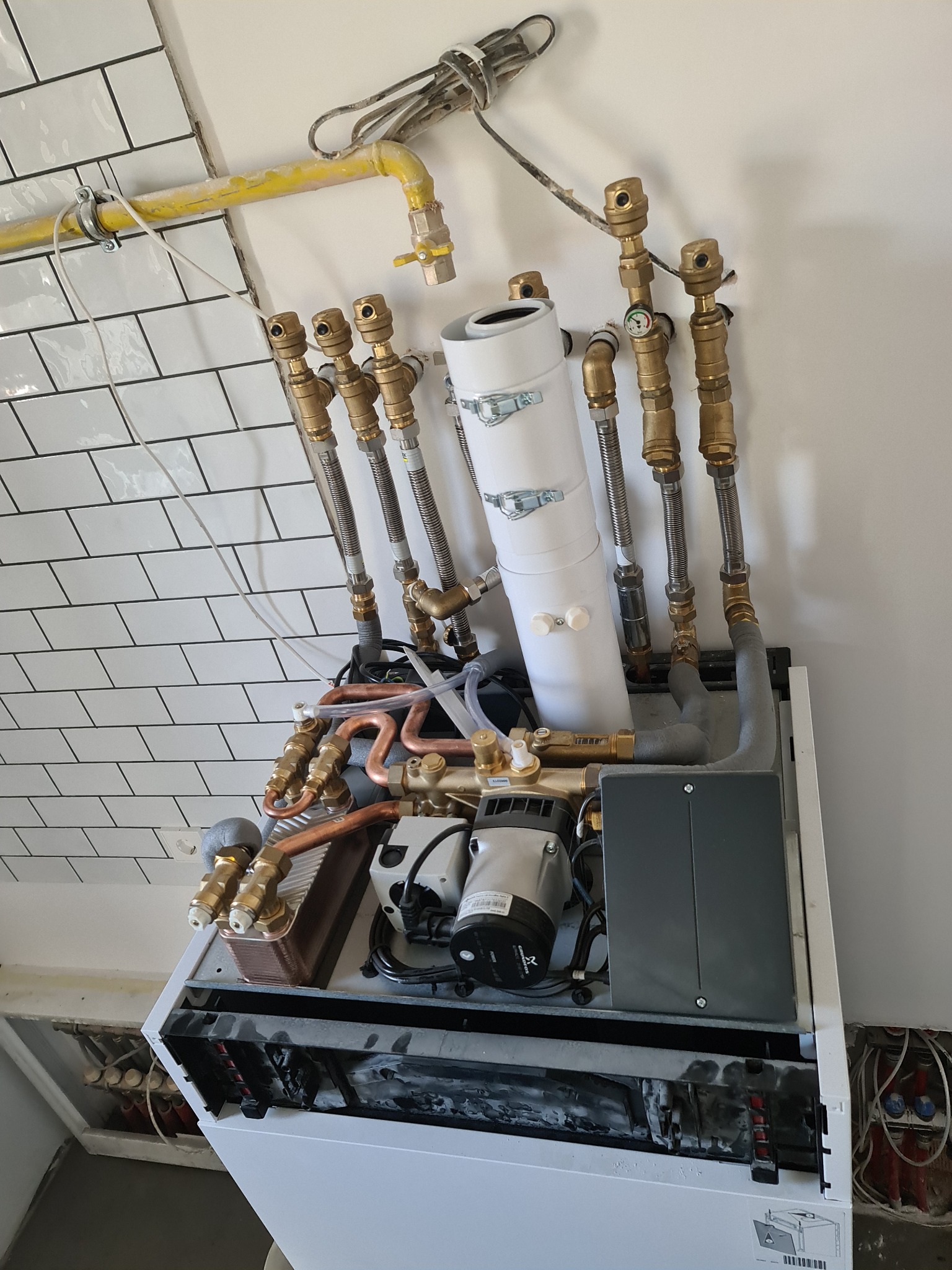 Otwarty piec grzewczy z widocznymi elementami instalacji, rurami miedzianymi i białym zbiornikiem, na tle białej ściany z częściowo ułożonymi białymi płytkami.