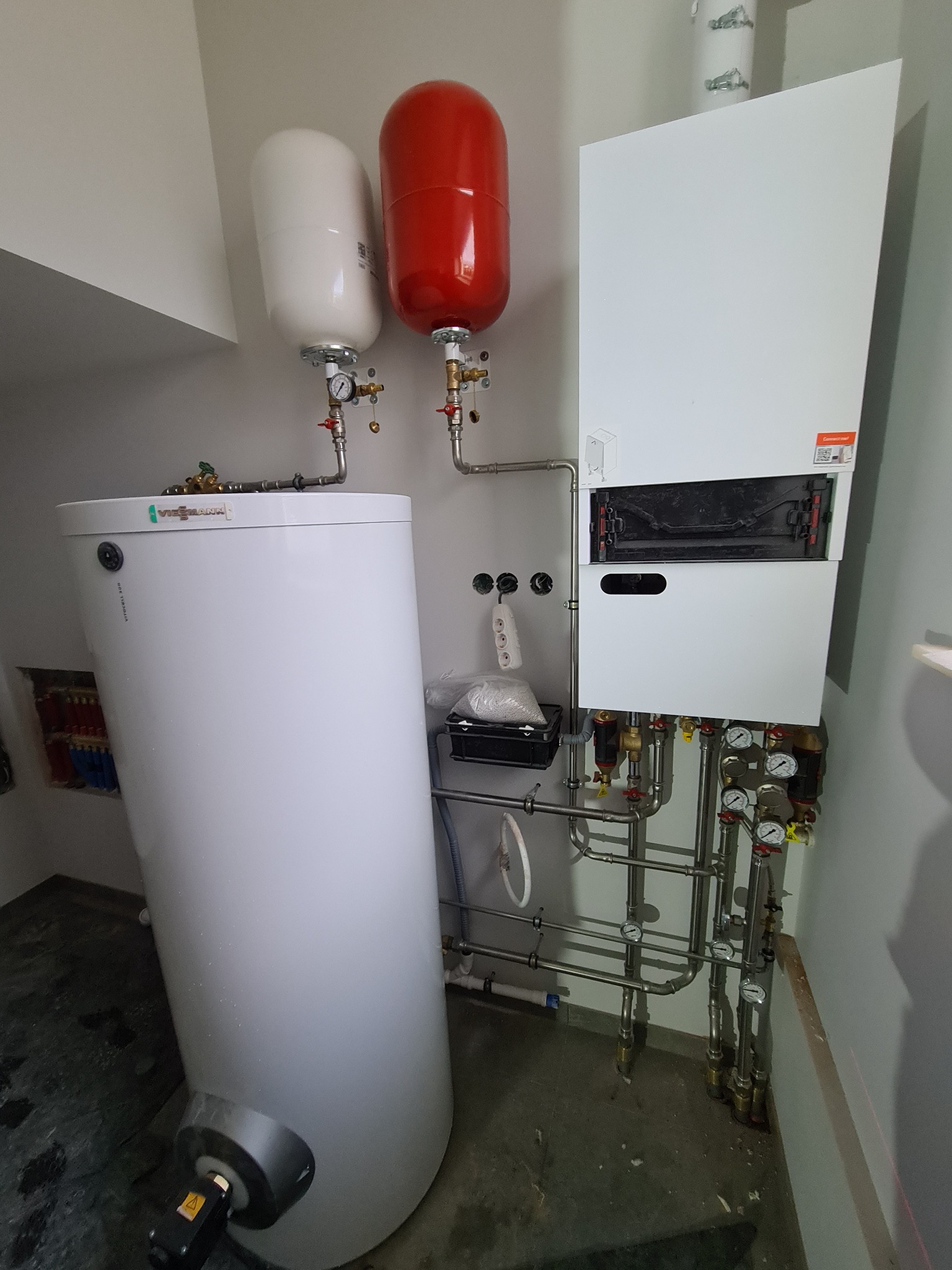 Nowoczesna kotłownia z białym zasobnikiem ciepłej wody, czerwonym i białym naczyniem przeponowym oraz piecem grzewczym, połączonych rurami z manometrami.
