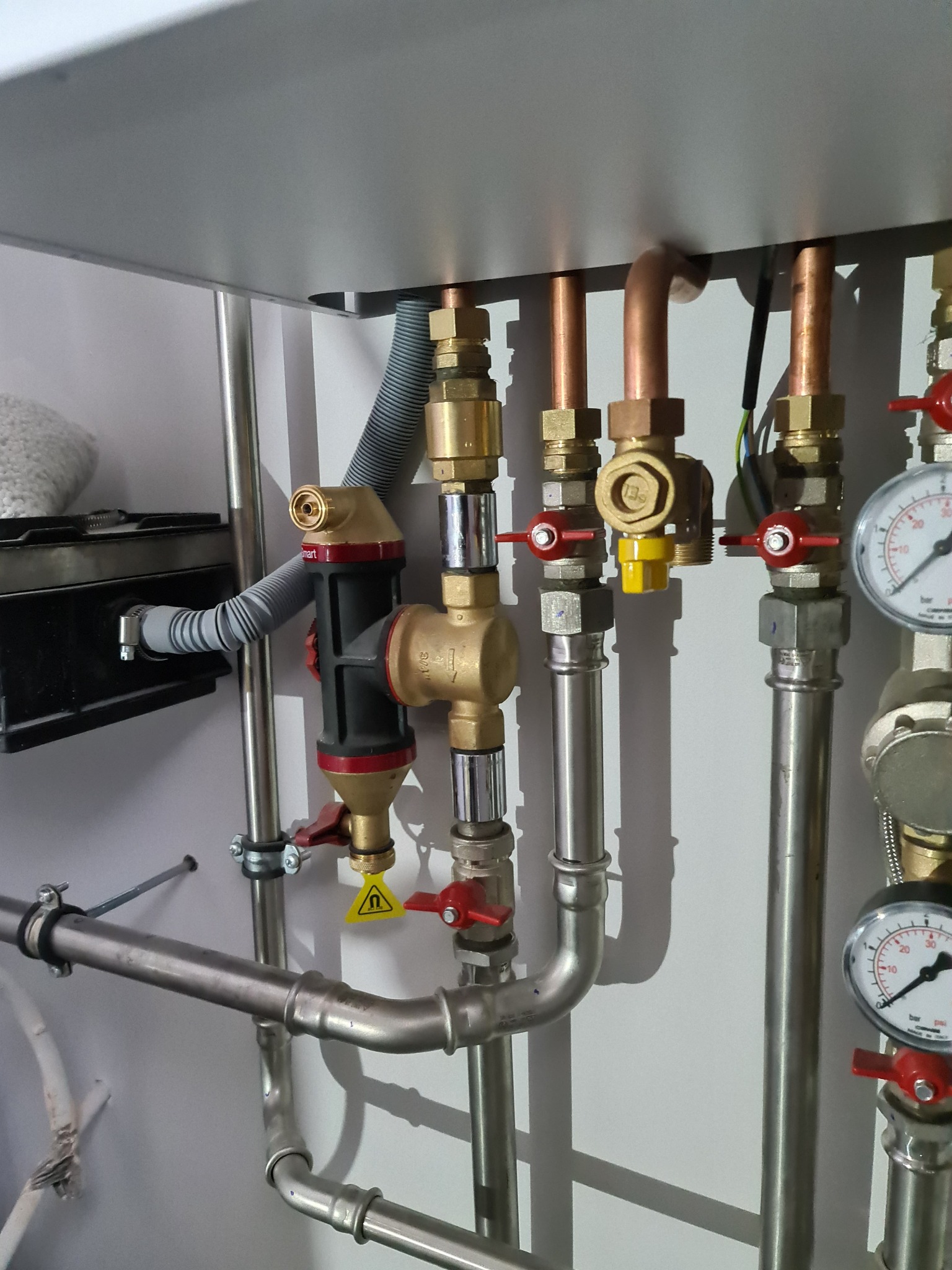 Szczegółowe ujęcie instalacji hydraulicznej z rurami miedzianymi i stalowymi, zaworami z czerwonymi uchwytami, manometrami i filtrem z żółtym symbolem ostrzegawczym na białej ścianie.