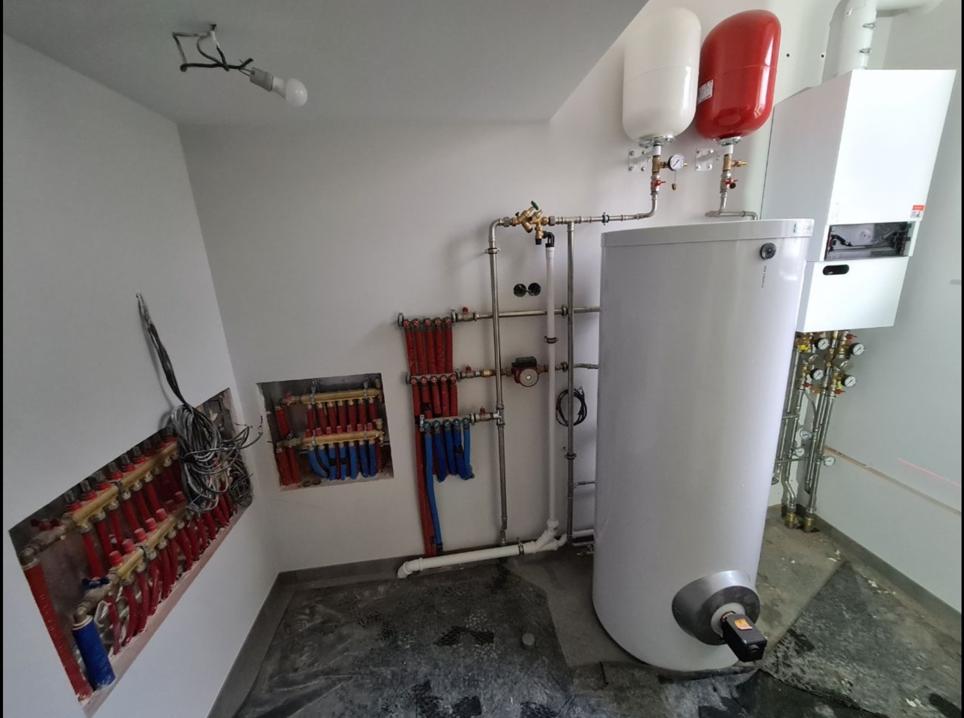 Kotłownia w trakcie instalacji: biały zasobnik ciepłej wody, czerwony i biały zbiornik wyrównawczy, rozdzielacze ogrzewania podłogowego w ścianie, liczne rury i zawory, surowy beton na podłodze...