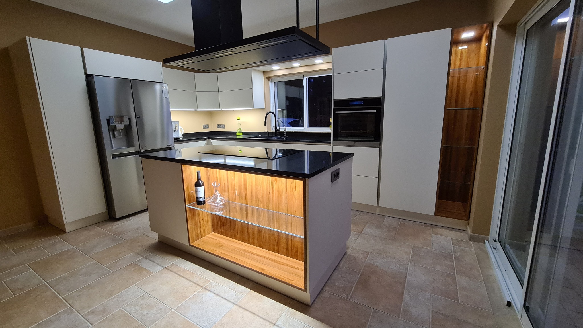 Nowoczesna kuchnia z wyspą, białymi szafkami, czarnym blatem i oświetleniem LED, z butelką wina i karafką na półce wyspy.