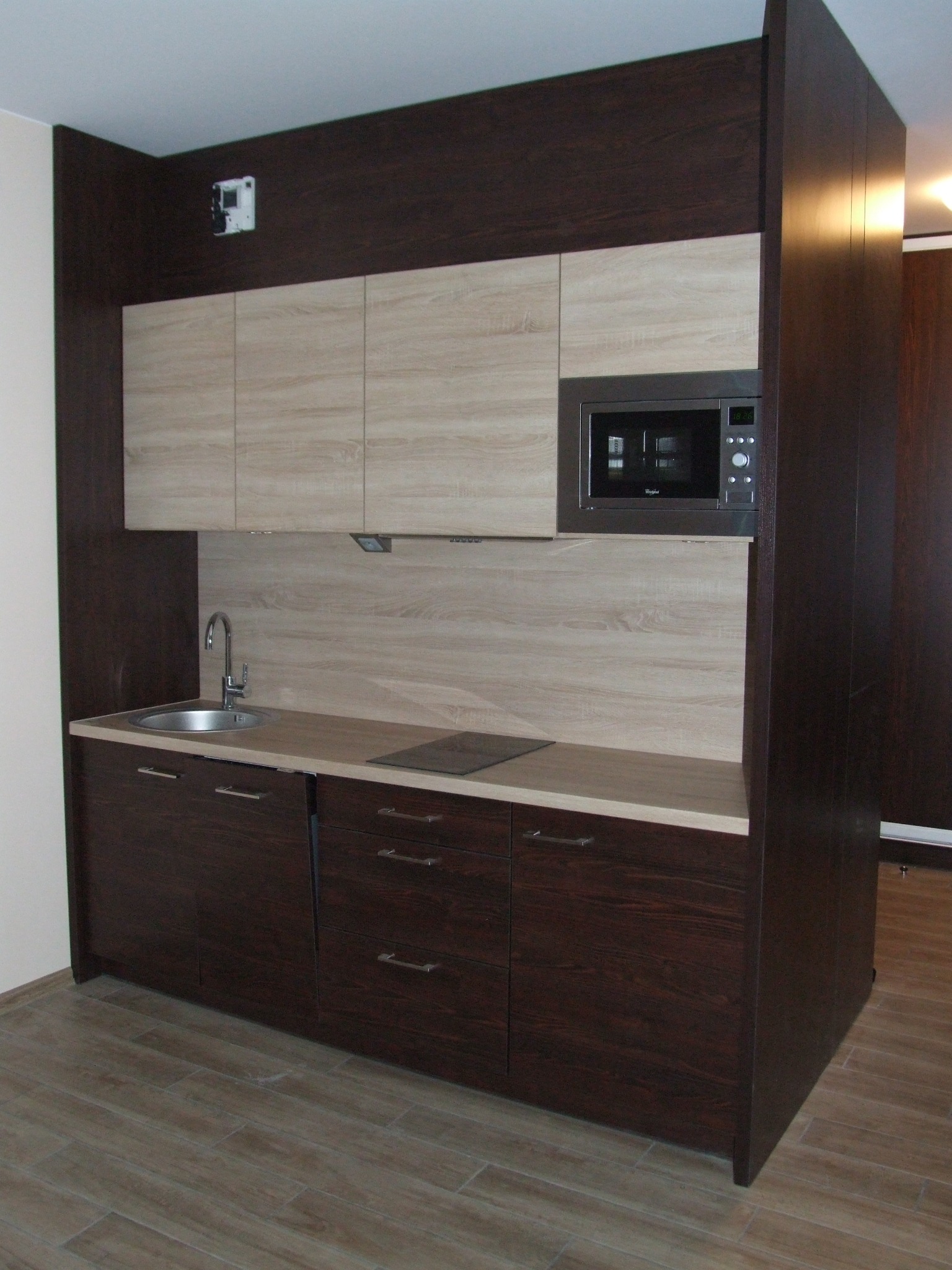 Kompaktowa zabudowa kuchenna z ciemnobrązowymi dolnymi szafkami i jasnymi górnymi szafkami z fakturą drewna, z wbudowaną mikrofalówką i płytą grzewczą, całość do sufitu, zlew po lewej stronie.