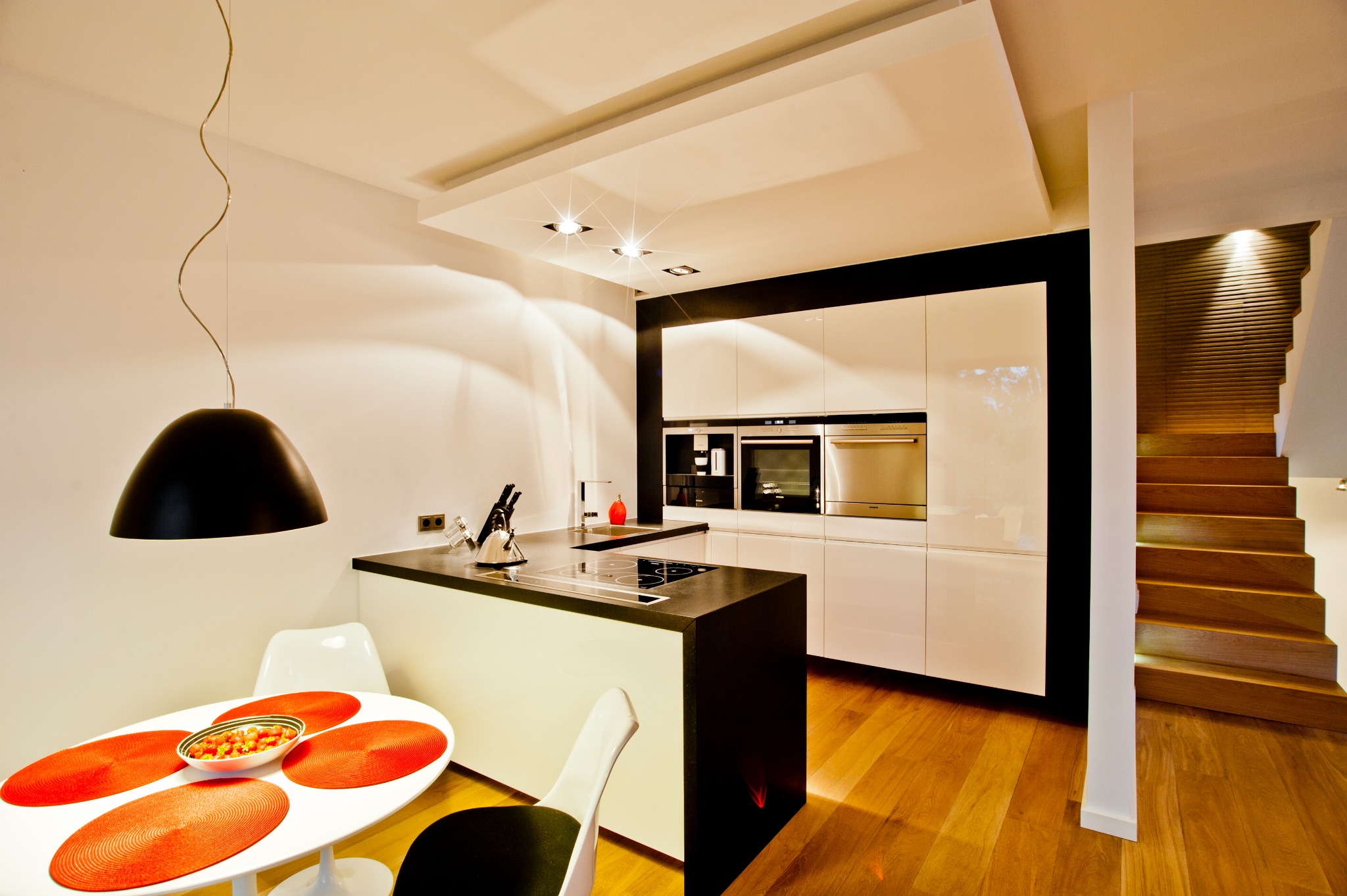 Nowoczesna kuchnia z białymi lakierowanymi szafkami, czarnym blatem wyspy i drewnianymi schodami w tle, oświetlona wiszącą lampą i halogenami.