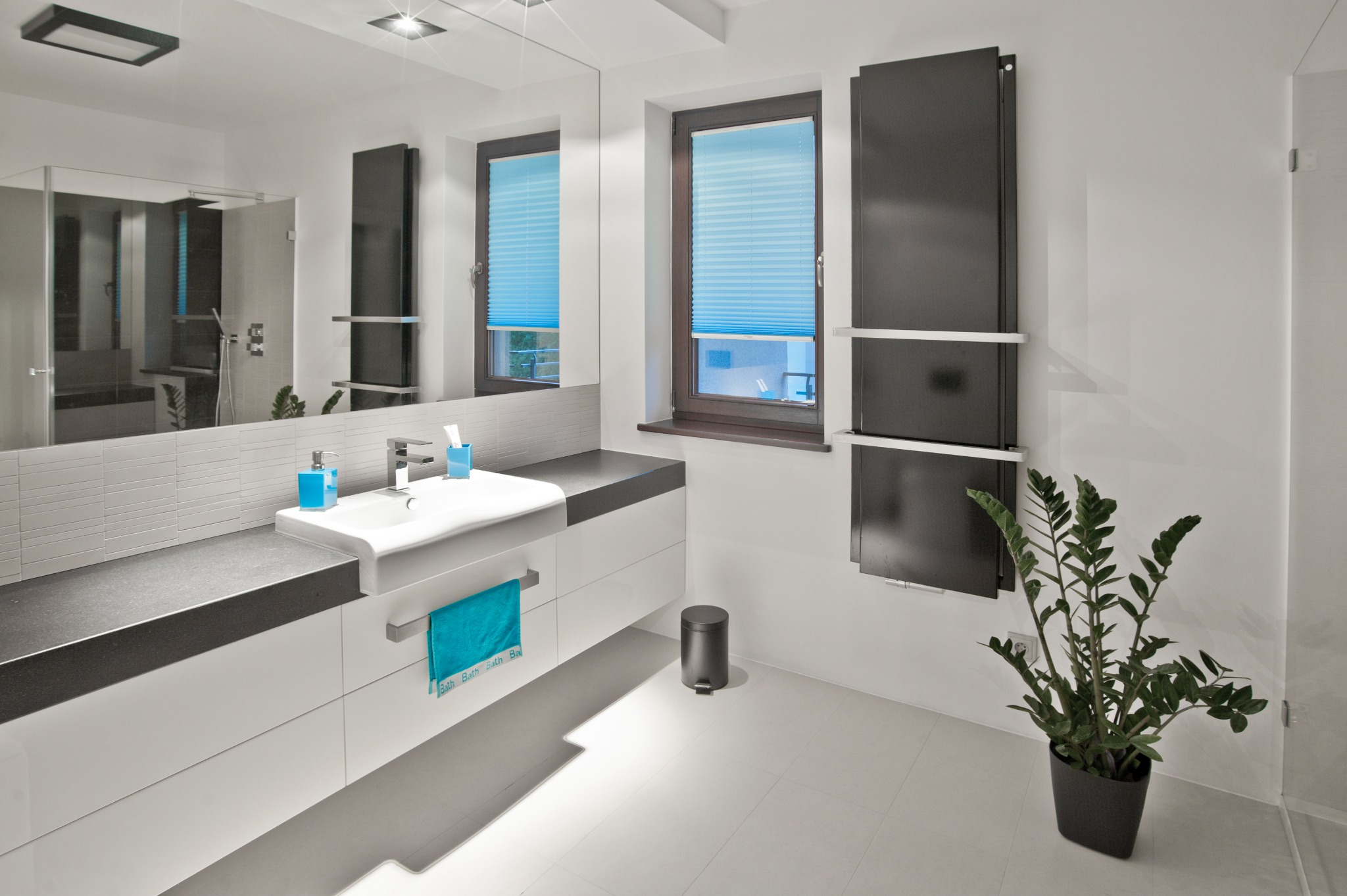 Nowoczesna łazienka z białymi szafkami pod umywalką z ciemnym blatem, duże lustro, grzejnik drabinkowy w kolorze antracytowym, okno z jasnoniebieskimi plisami i roślina doniczkowa.
