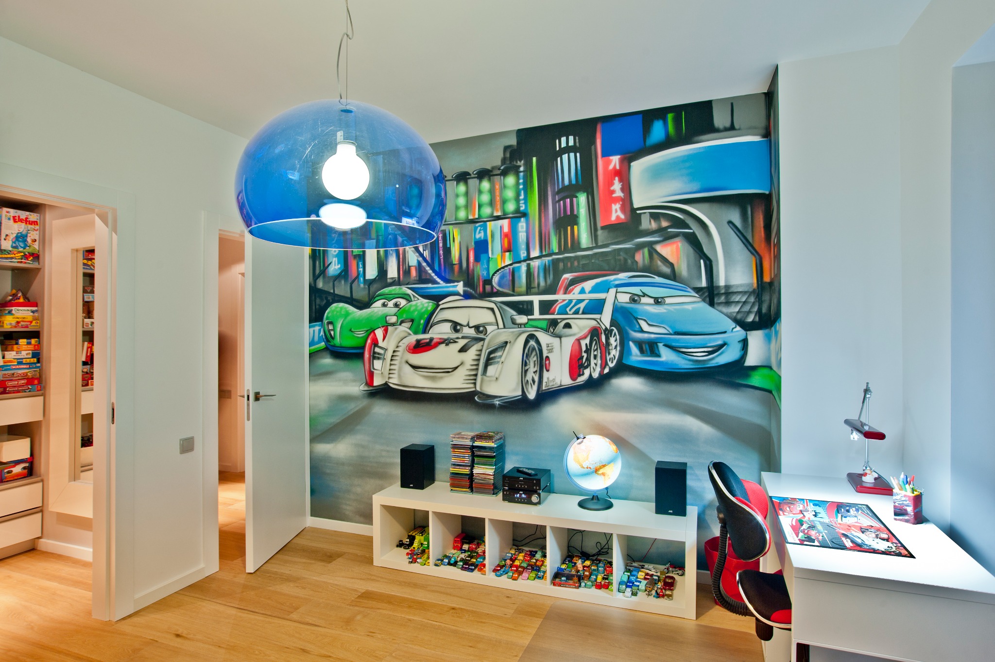 Pokój dziecięcy z muralem przedstawiającym postacie z filmu animowanego 'Auta', białe meble, drewniana podłoga i duża, błękitna lampa wisząca.