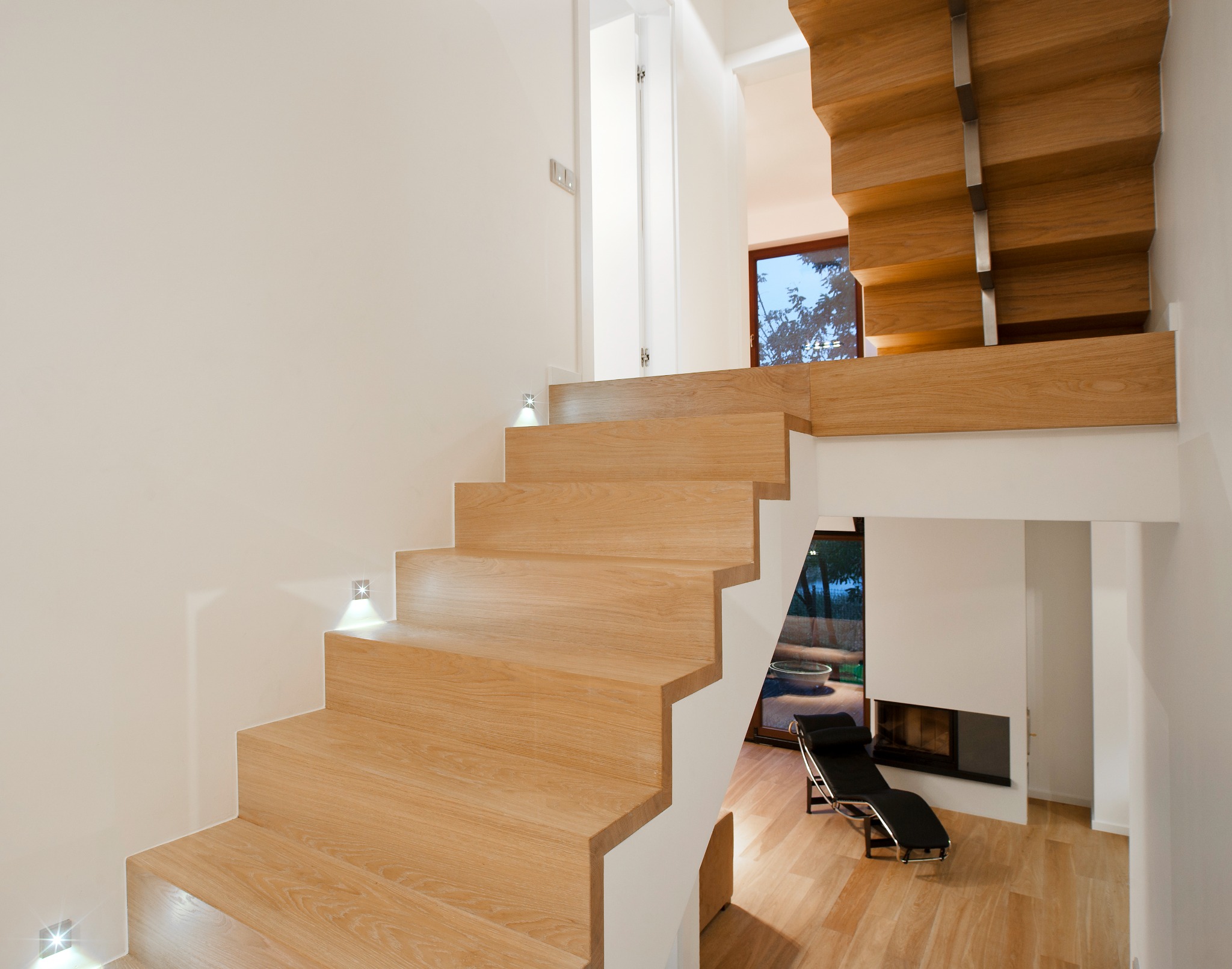 Nowoczesne drewniane schody z podświetleniem LED w minimalistycznym wnętrzu, widok na salon z kominkiem i leżanką.