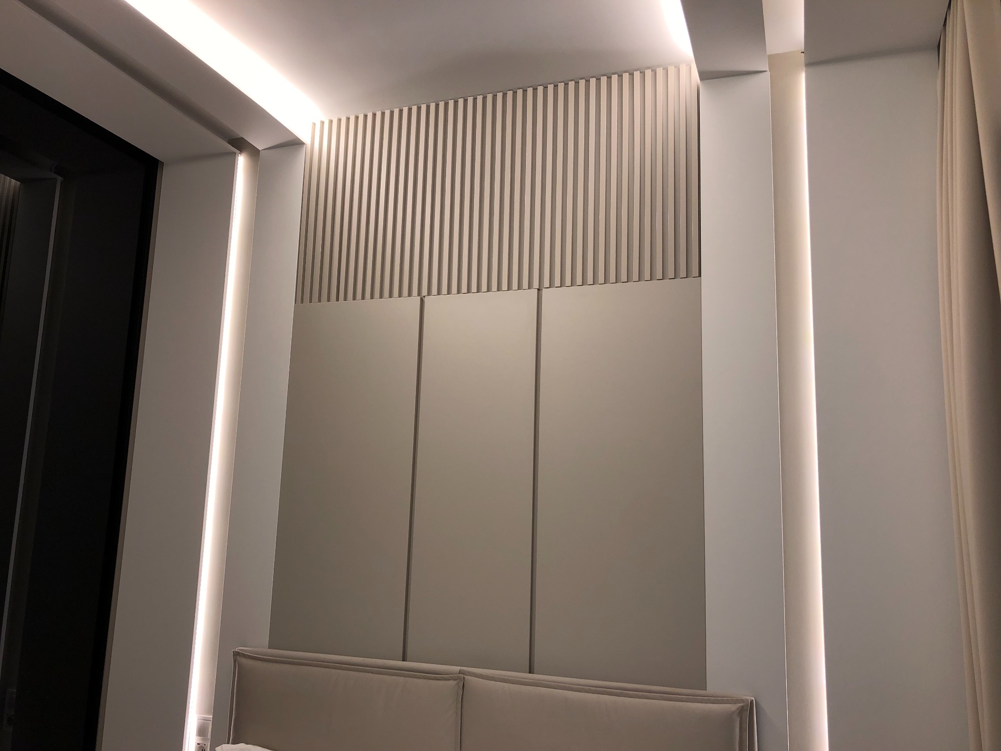 Nowoczesna szafa wnękowa z frontami w kolorze złamanej bieli, zintegrowana z panelem dekoracyjnym z pionowych lameli i oświetleniem LED wzdłuż krawędzi, widoczny fragment wezgłowia łóżka.