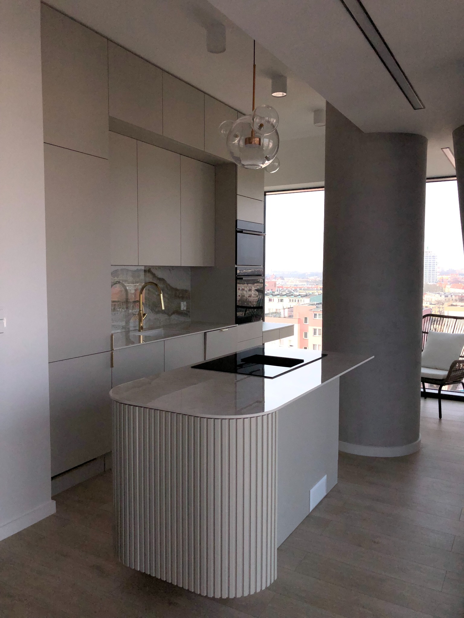 Nowoczesna kuchnia z wyspą, jasnymi szafkami i złotą armaturą na tle panoramy miasta widocznej przez duże okno.