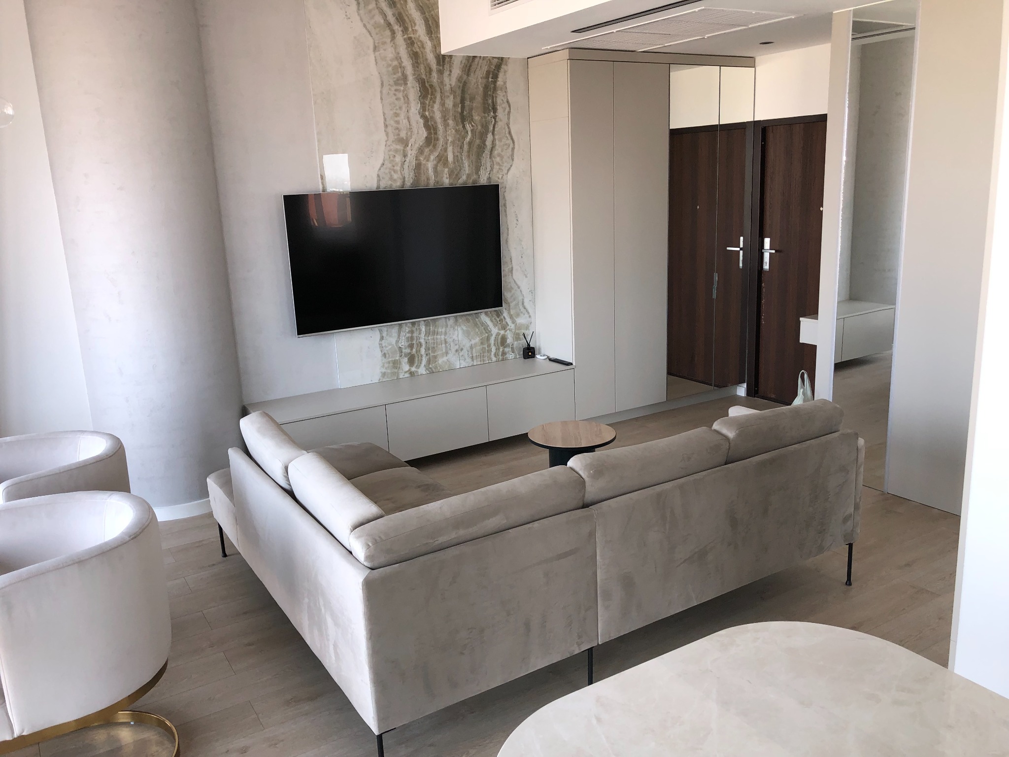 Nowoczesny salon z narożną sofą w odcieniach beżu, minimalistyczną szafką RTV pod telewizorem, ścianą z imitacją marmuru i widocznymi drzwiami do szafy w zabudowie.