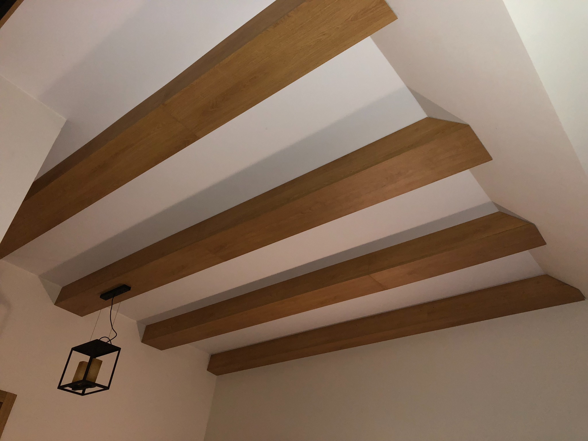 Drewniane belki stropowe w jasnym pomieszczeniu, z wiszącą, czarną, geometryczną lampą z imitacją świec.