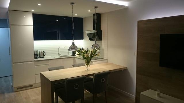 Nowoczesna kuchnia w Przecławiu z białymi frontami szafek, ciemnoniebieskim panelem nad blatem, wysuwanym drewnianym stołem jadalnym i wiszącymi lampami w stylu industrialnym.