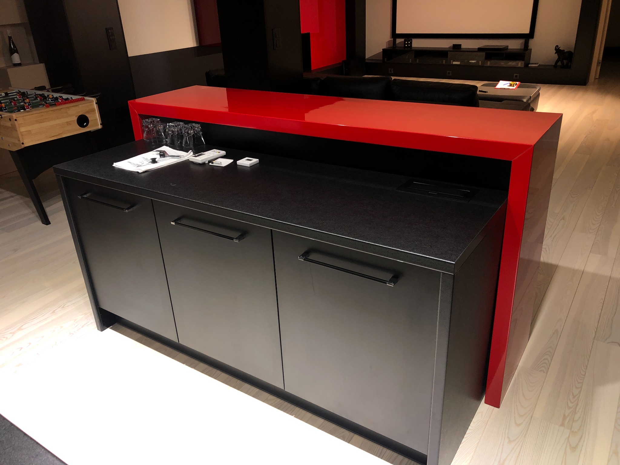 Nowoczesna zabudowa meblowa z czarnymi frontami i czerwonym blatem, uzupełniona szklankami i notatkami na blacie, w tle stół do gry w piłkarzyki i ekran projekcyjny.