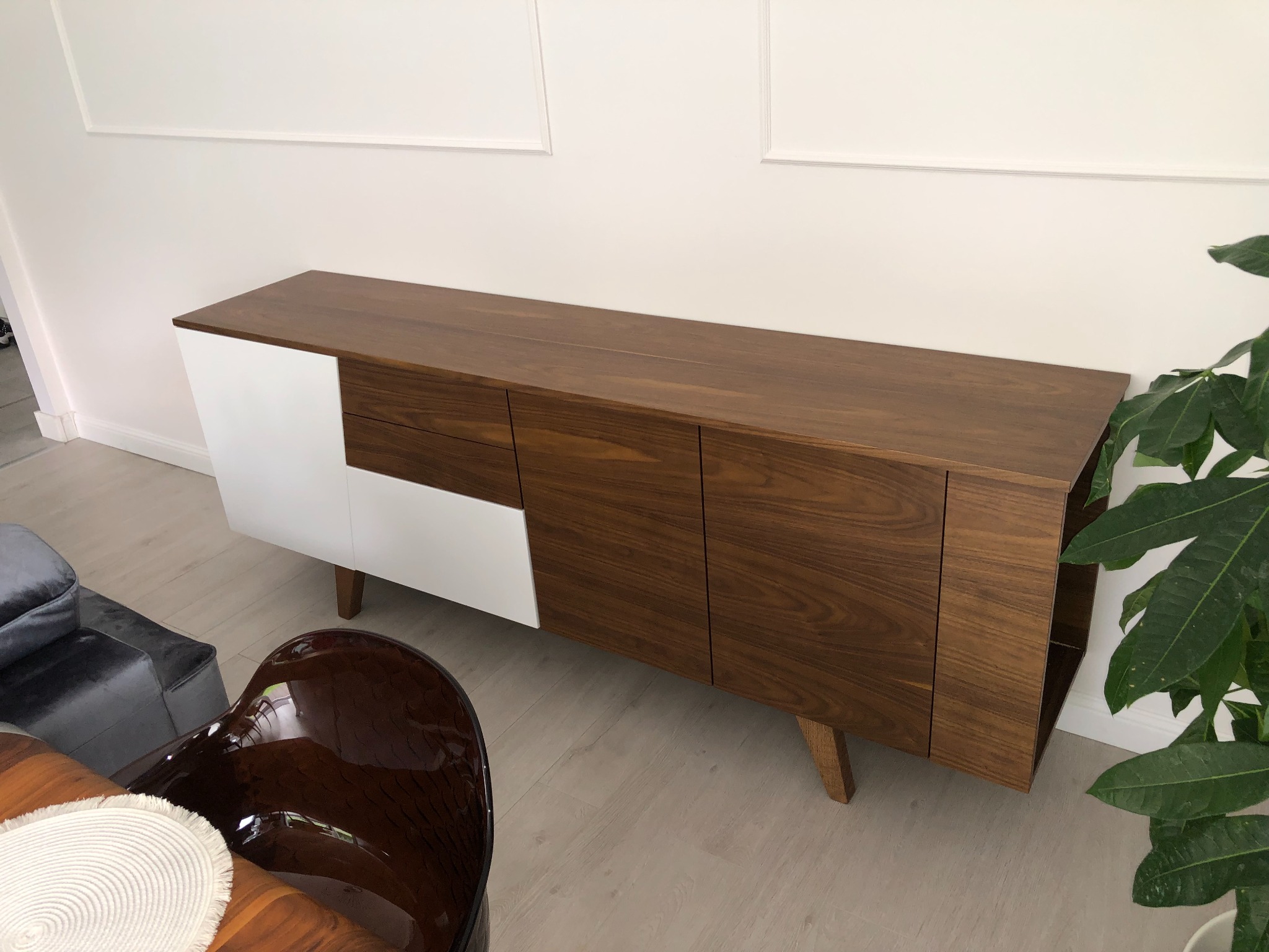 Nowoczesna komoda z drewna w odcieniach brązu i bieli, z minimalistycznym designem i widocznymi słojami drewna, ustawiona w jasnym wnętrzu obok krzesła z brązowego tworzywa i rośliny doniczkowej.