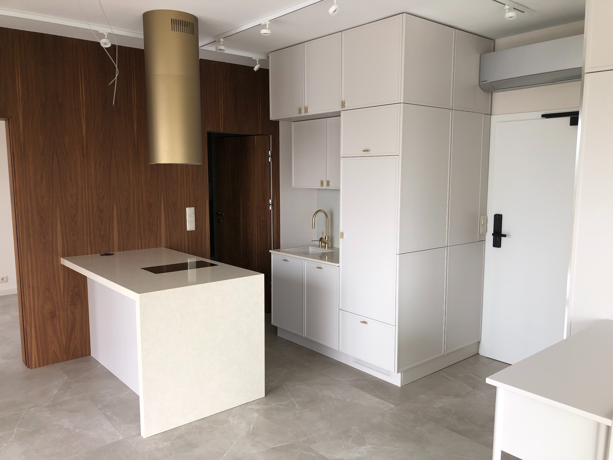 Nowoczesna kuchnia z białymi szafkami na wymiar, złotą armaturą i wyspą z blatem kwarcowym, uzupełniona o drewnianą ścianę i złotą tubę wentylacyjną.