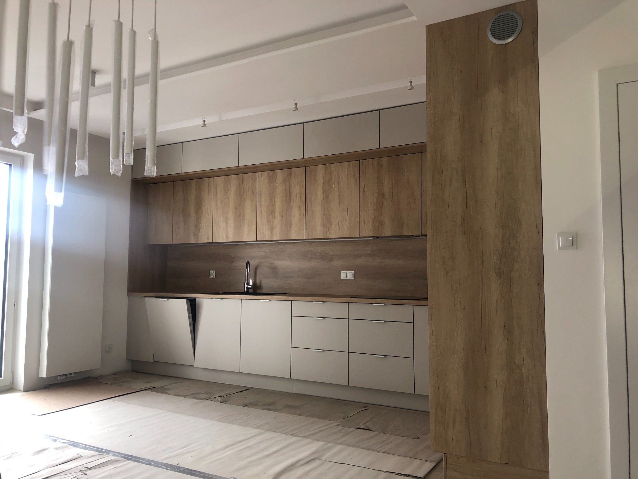Nowoczesna kuchnia z drewnianymi i szarymi frontami, jeszcze w trakcie wykańczania, z folią ochronną na podłodze i lampami w osłonach.