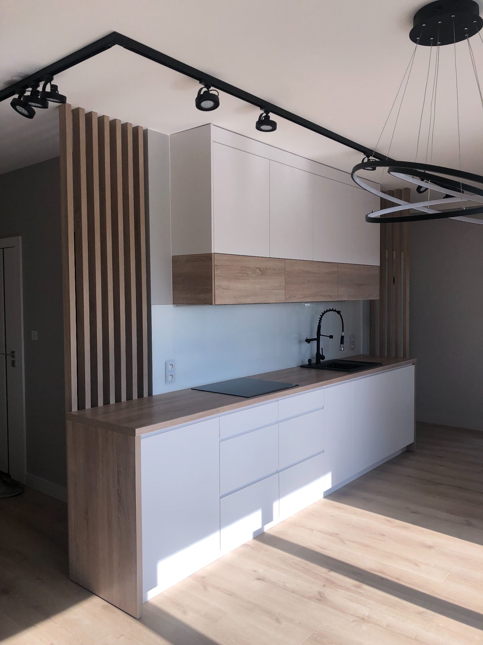 Nowoczesna kuchnia z białymi szafkami i drewnianymi blatami, czarną armaturą i oświetleniem punktowym, oddzielona od reszty pomieszczenia drewnianymi lamelami.