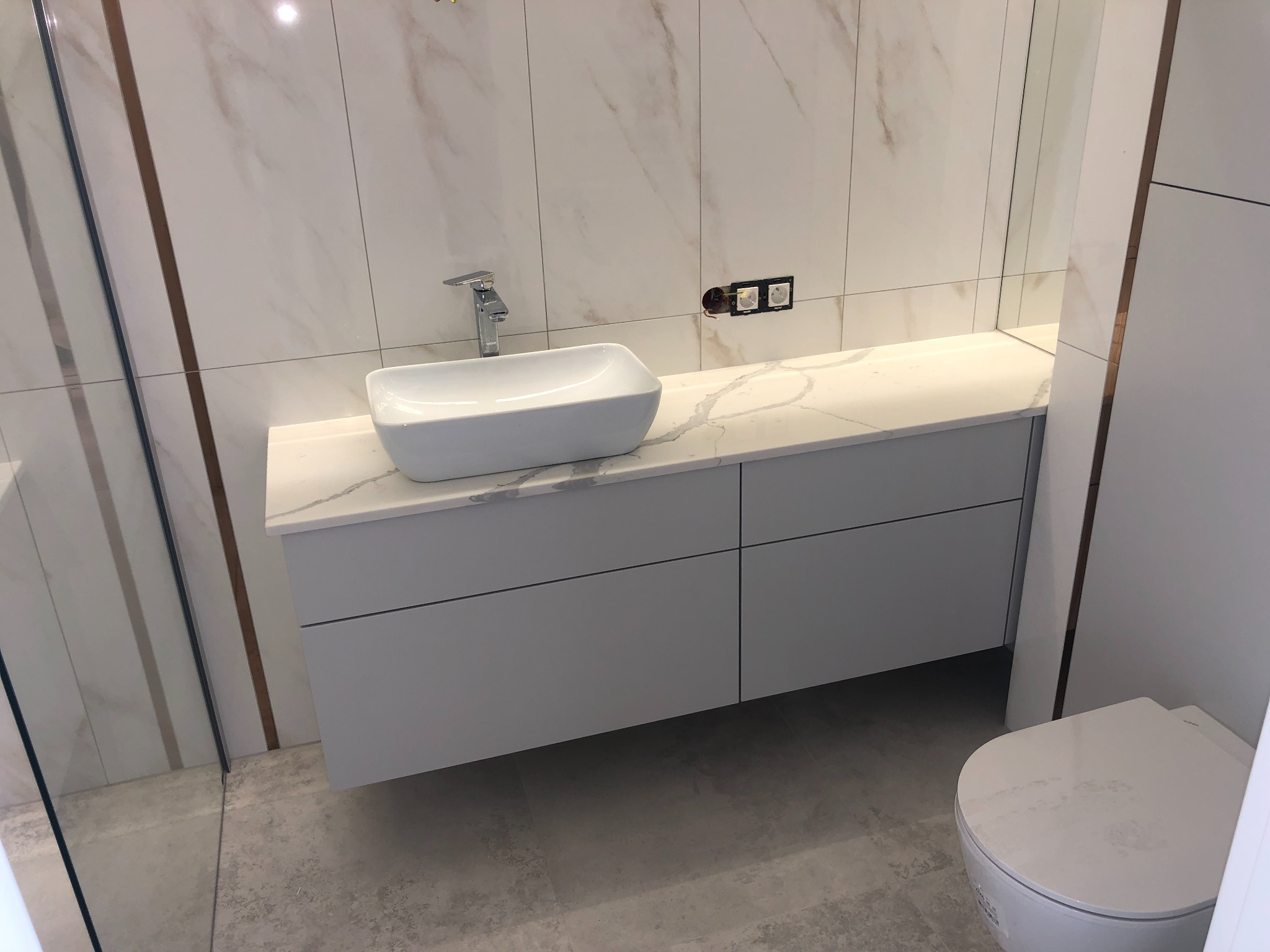 Nowoczesna łazienka z białą, prostokątną umywalką nablatową, szafką z szufladami bez uchwytów, marmurowymi płytkami ściennymi z brązowymi żyłkami i widocznymi gniazdkami elektrycznymi.