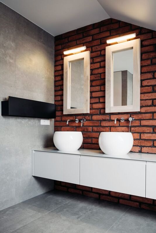 Nowoczesna łazienka z dwoma okrągłymi umywalkami nablatowymi, białymi szafkami pod umywalką, ceglaną ścianą, dwoma lustrami w jasnych drewnianych ramach i podłużnymi lampami nad nimi.