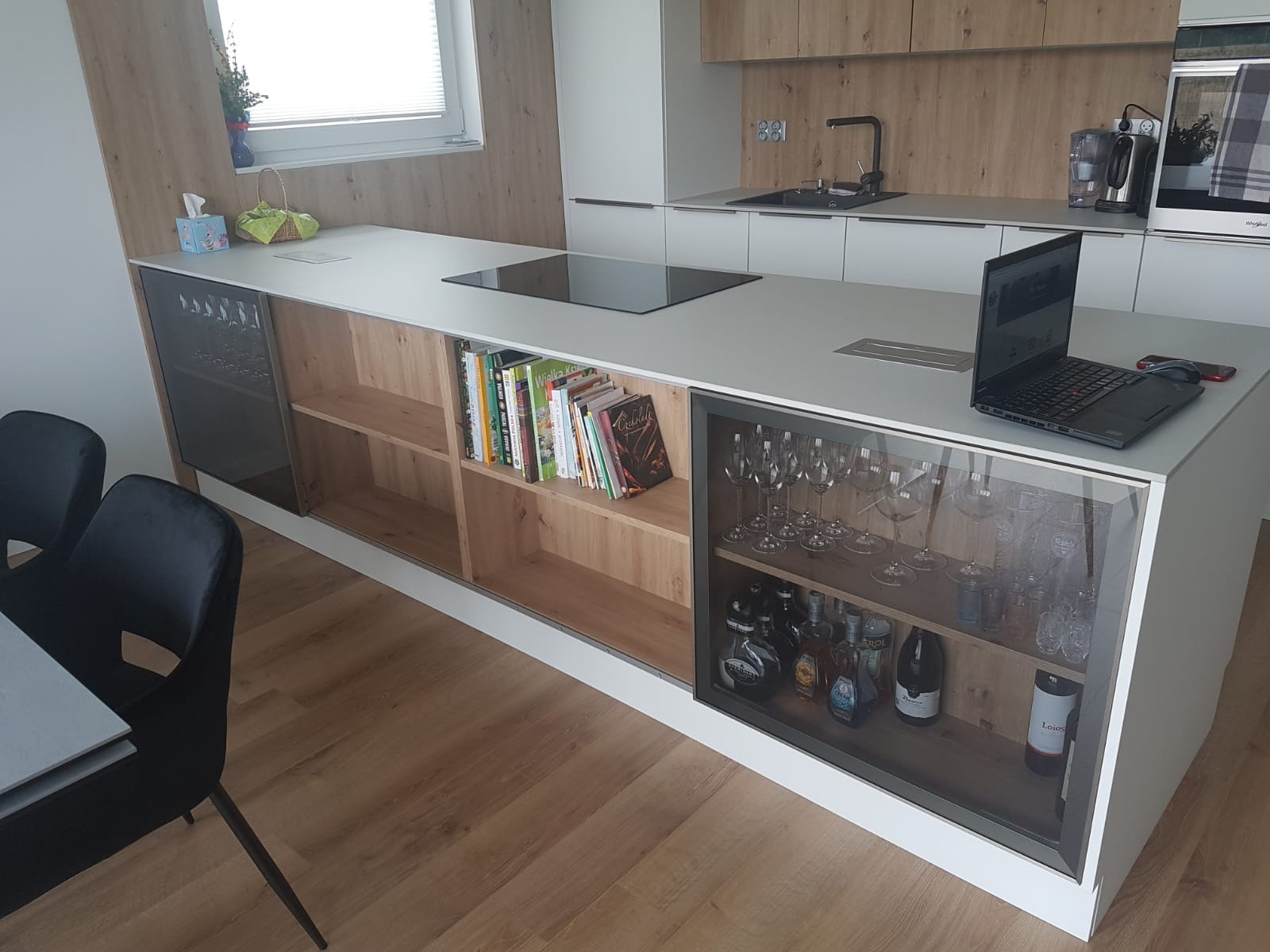 Wyspa kuchenna z białym blatem i zabudową z drewnianymi frontami, wyposażona w szklane witryny prezentujące kolekcję kieliszków i alkoholi, na blacie laptop i płyta indukcyjna.