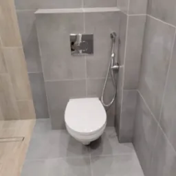 Nowoczesna toaleta podwieszana z chromowanym przyciskiem spłukującym i bidetką w szarej łazience wykończonej płytkami w jednolitym odcieniu.