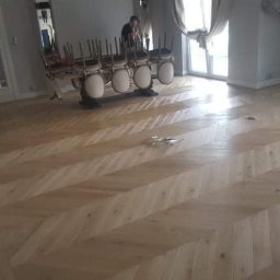 MKParkiety - Jasny, ułożony w jodełkę parkiet w przestronnym salonie z dużymi oknami i krzesłami ustawionymi na stole w tle.