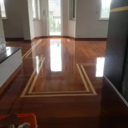 Wypolerowany parkiet z ciemnego drewna z dekoracyjną jasną obwódką, widok na przestronny pokój z oknami i drzwiami balkonowymi, wiadro z narzędziami w rogu.