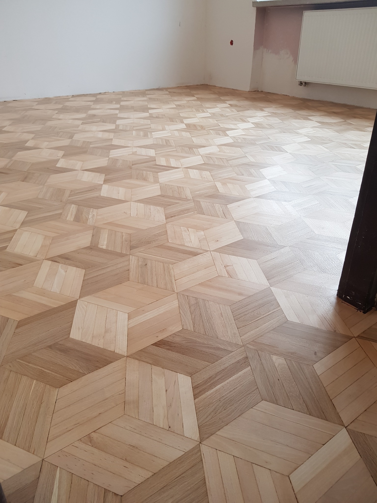 Ułożony parkiet w geometryczny wzór 3D, jasne drewno, widok z góry, częściowo wykończone ściany w tle.