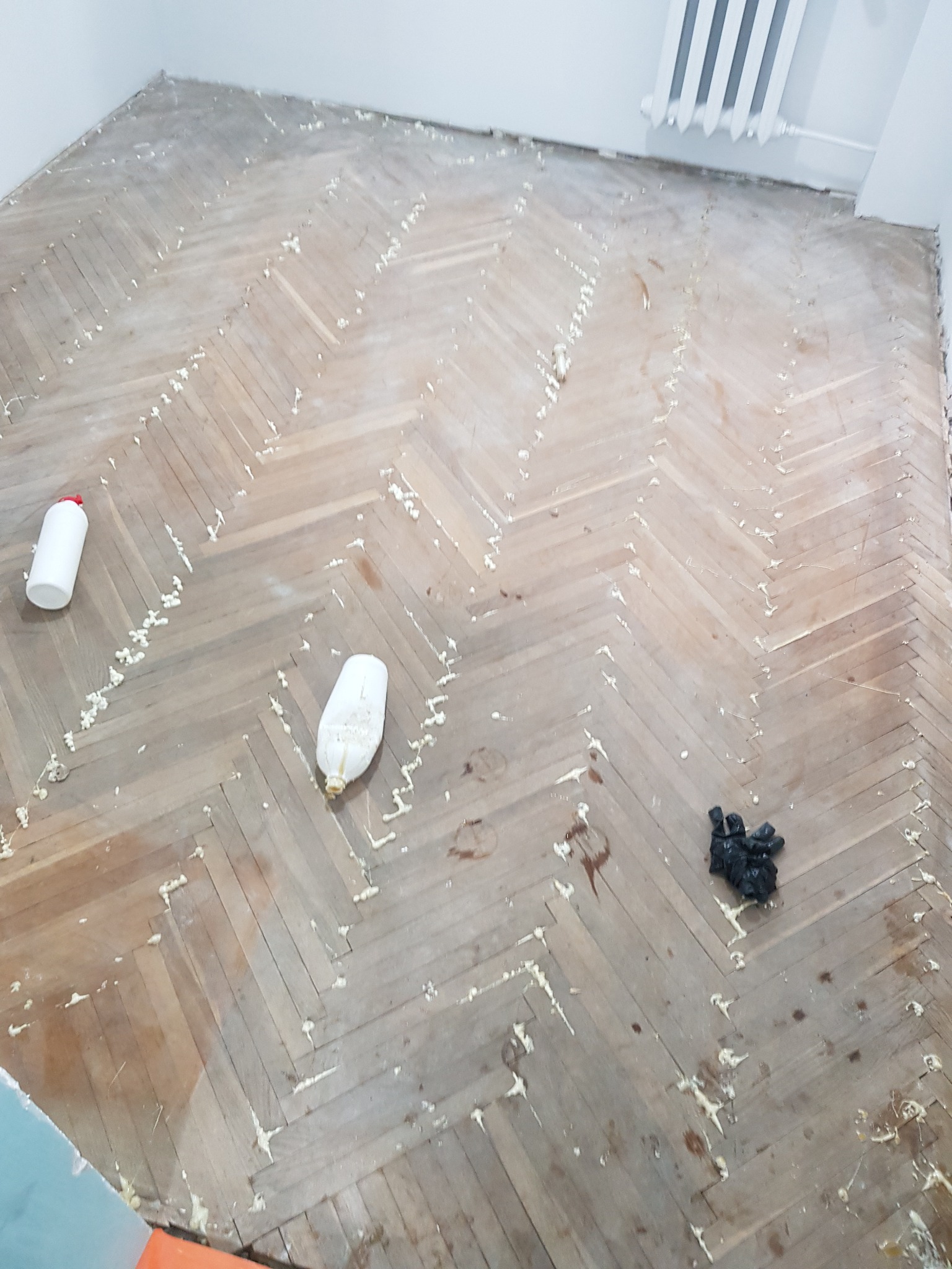 Stary parkiet w jodełkę, częściowo oczyszczony z kleju, z widocznymi butelkami po preparatach i workiem na śmieci w rogu pokoju.