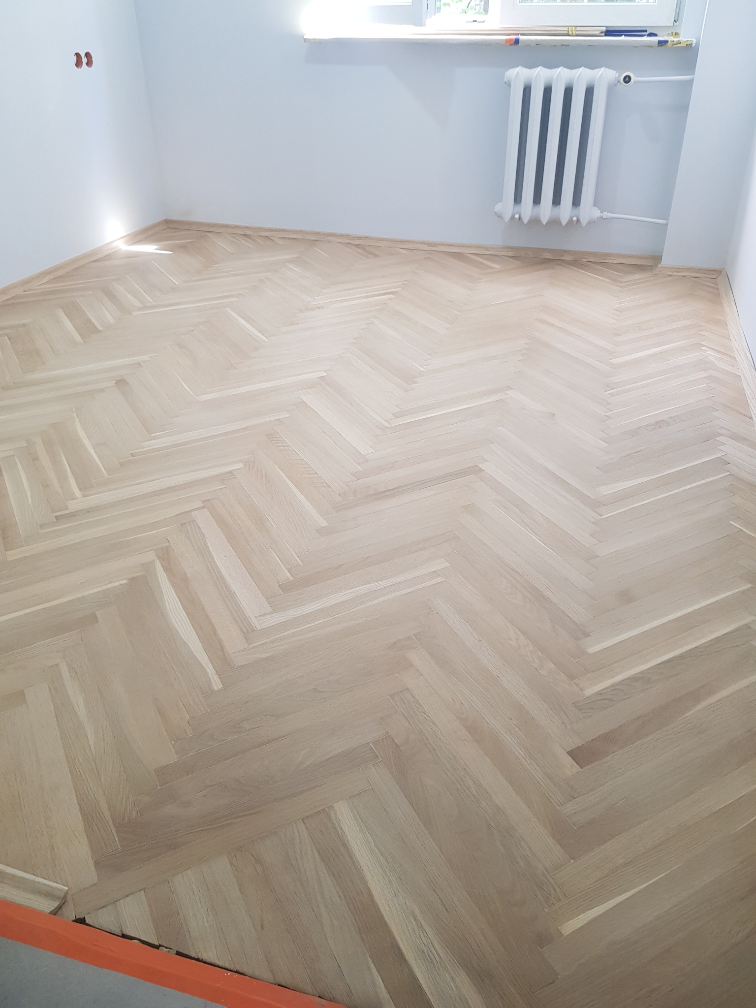 Jasny, nowy parkiet ułożony we wzór jodełki francuskiej w pustym pokoju z grzejnikiem i listwami przypodłogowymi.