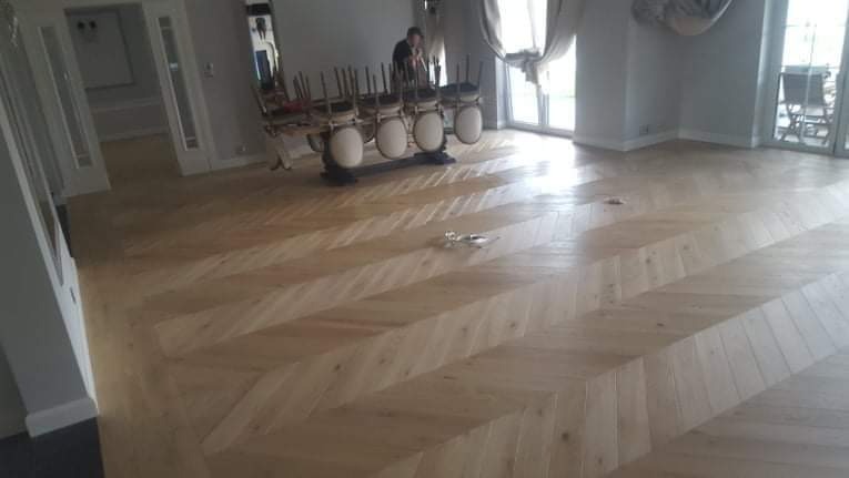 Jasny, ułożony w jodełkę parkiet w przestronnym salonie z dużymi oknami i krzesłami ustawionymi na stole w tle.