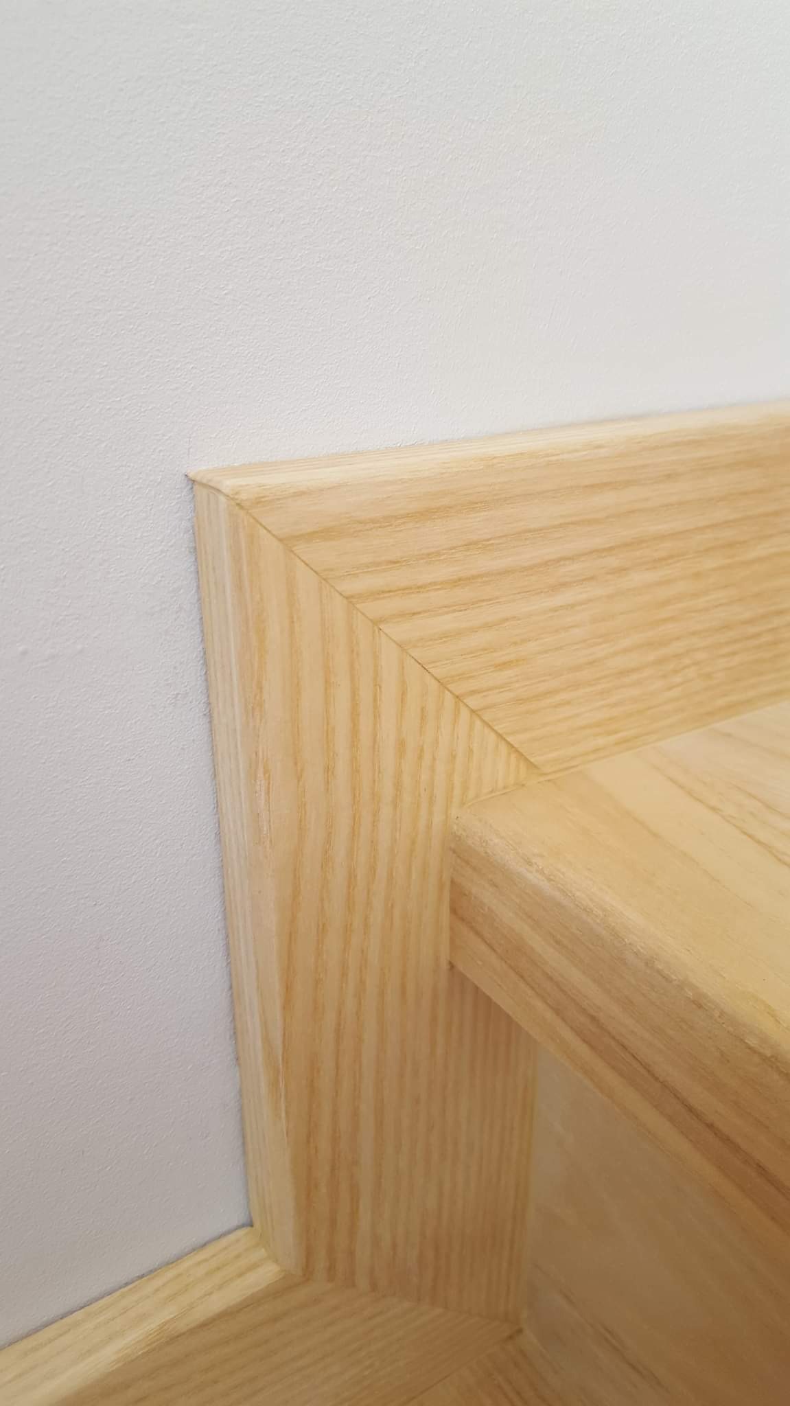 Fragment drewnianych schodów z listwą przypodłogową, widoczny narożnik stopnia i pionowa część, jasne drewno, biała ściana w tle.