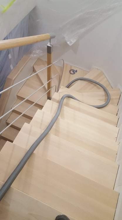 Jasne, drewniane schody z metalową balustradą częściowo zabezpieczone folią malarską, z widocznym wężem od odkurzacza przemysłowego podczas prac wykończeniowych.