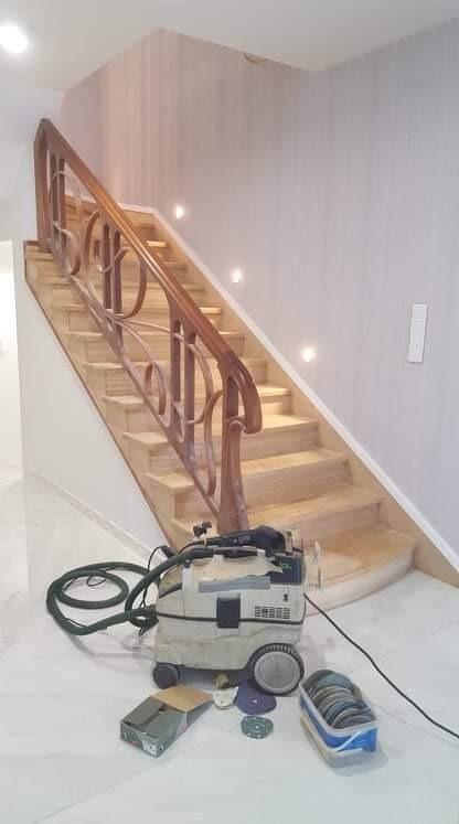 Drewniane schody z ozdobną balustradą w trakcie renowacji, widoczny odkurzacz przemysłowy i akcesoria do szlifowania, oświetlenie punktowe w ścianie.