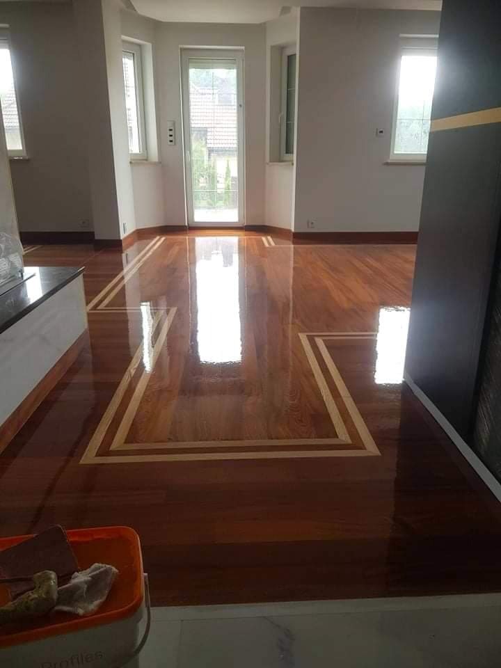 Wypolerowany parkiet z ciemnego drewna z dekoracyjną jasną obwódką, widok na przestronny pokój z oknami i drzwiami balkonowymi, wiadro z narzędziami w rogu.