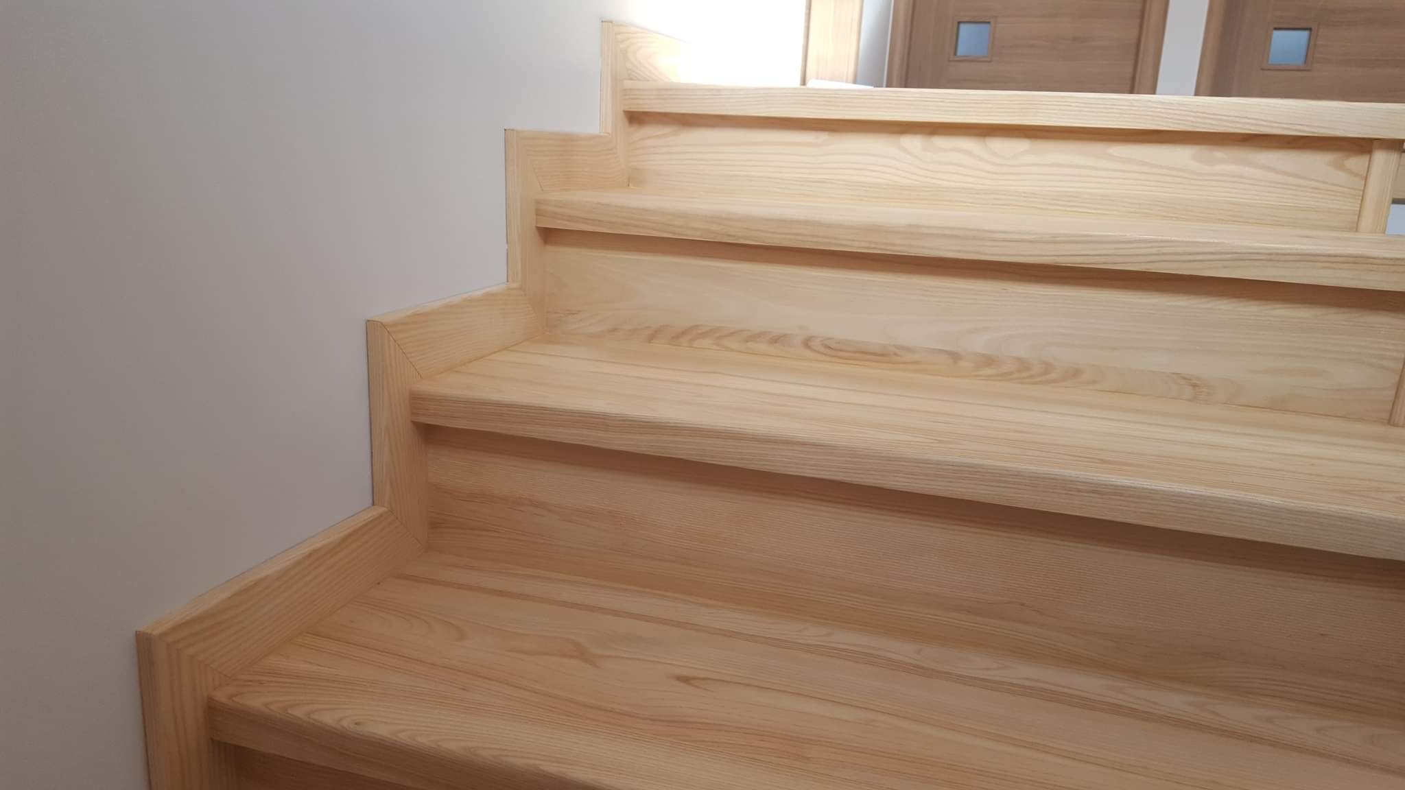 Jasne, drewniane schody z widocznym usłojeniem drewna, z dwiema parami drzwi w tle.