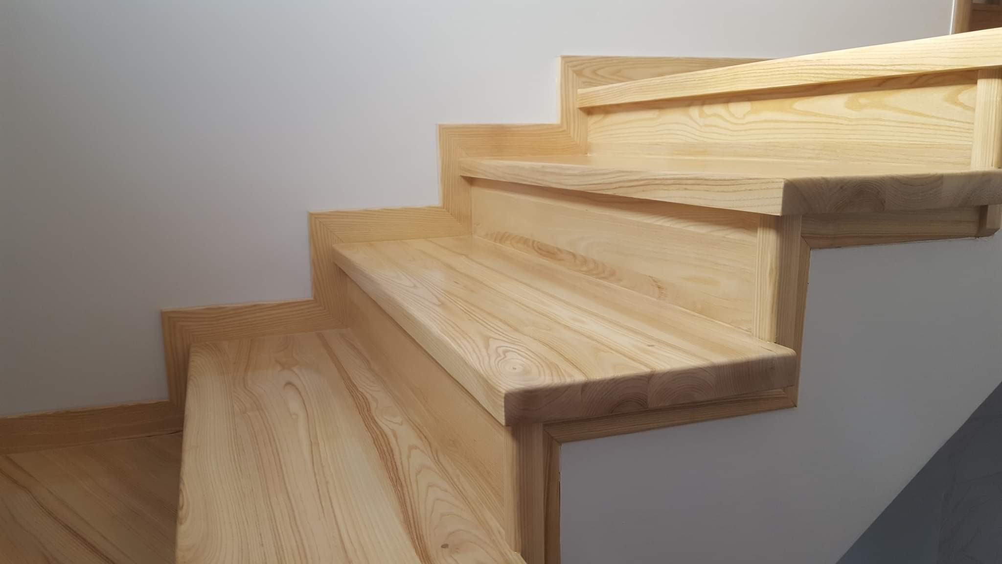 Jasne, drewniane schody z wyraźnym rysunkiem słojów, zamontowane przy białej ścianie, z widocznymi detalami wykończenia krawędzi.