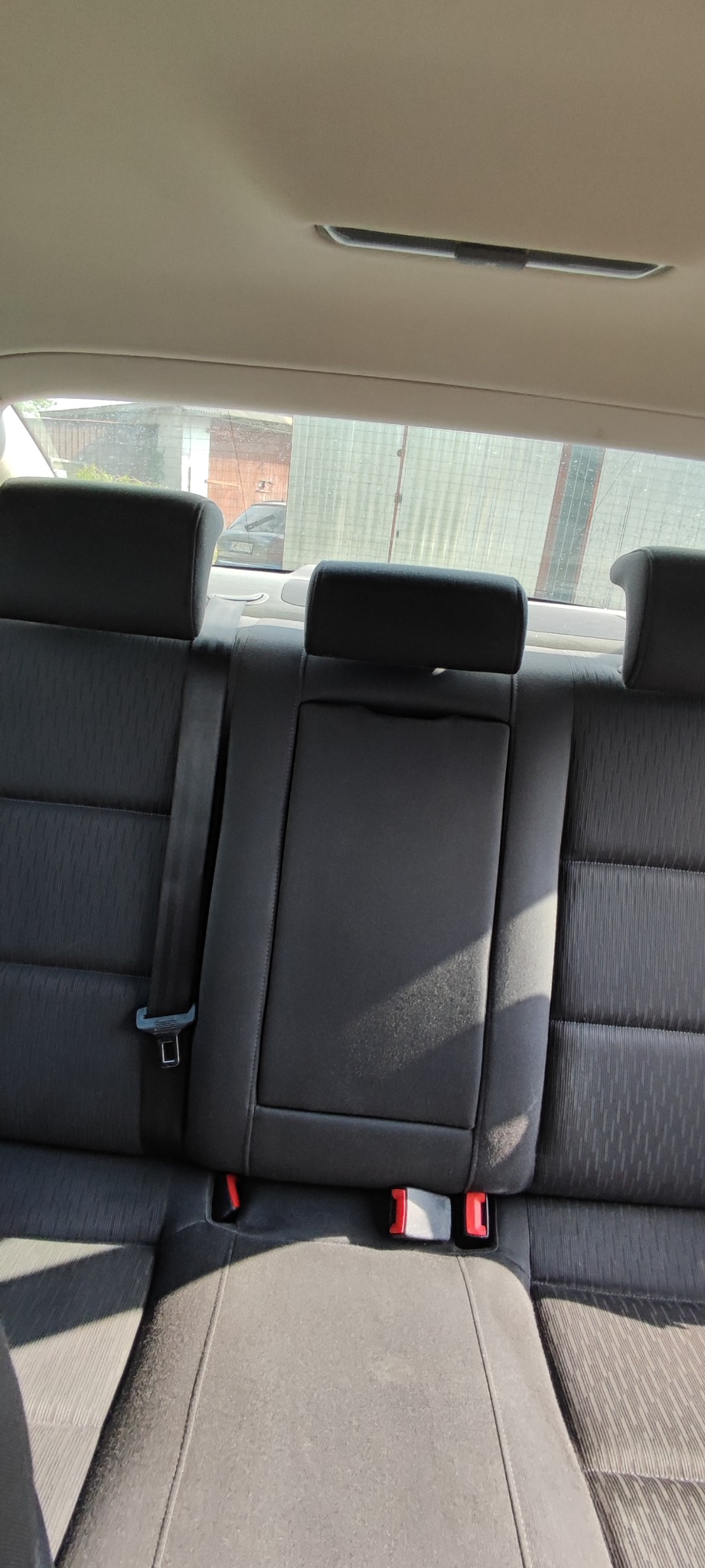 Wnętrze samochodu z widoczną tylną kanapą z materiałową tapicerką w odcieniach szarości i czerni, pasami bezpieczeństwa i zagłówkami, widok z tyłu pojazdu na zewnątrz.