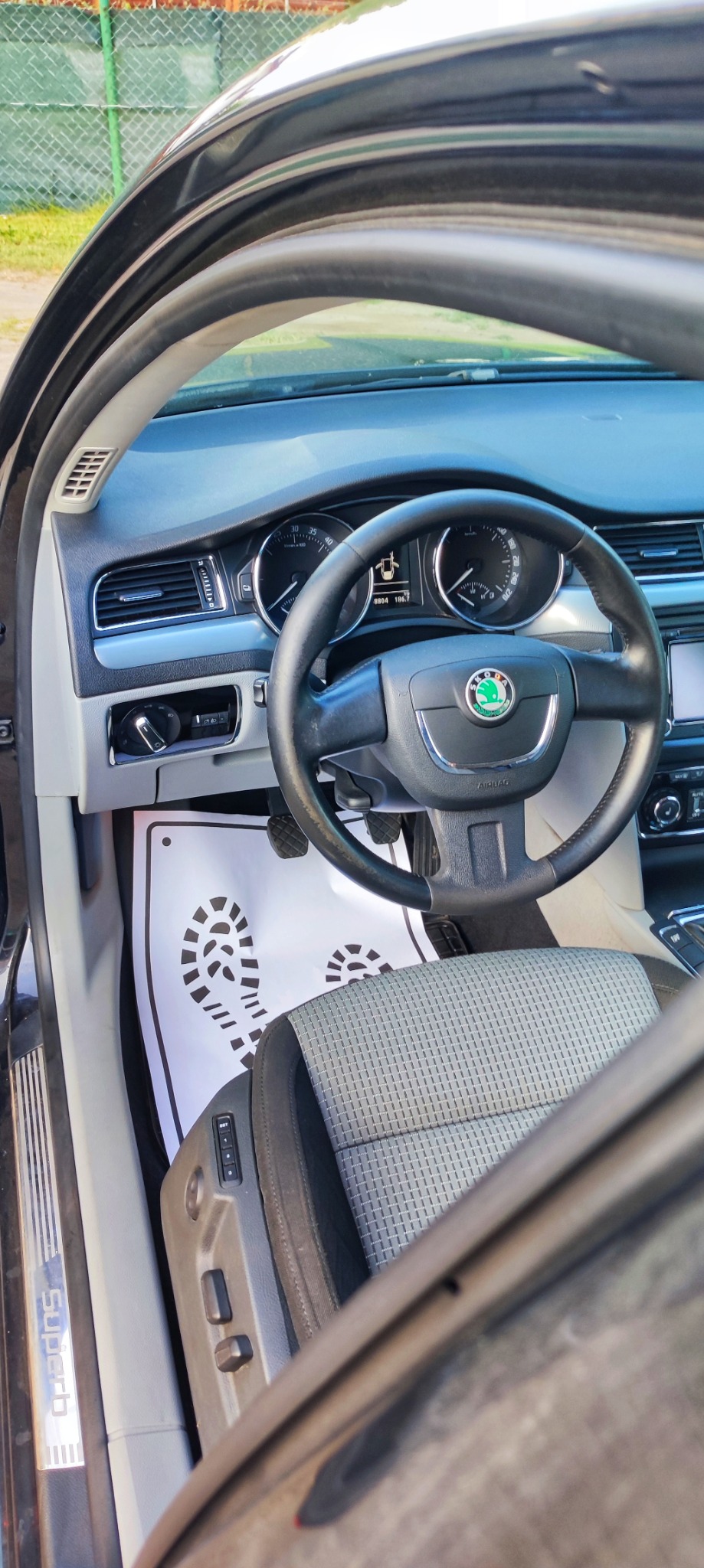 Wnętrze samochodu Skoda Superb z widoczną kierownicą, deską rozdzielczą i siedzeniem kierowcy, na dywaniku ochronny papier z nadrukiem śladów stóp.