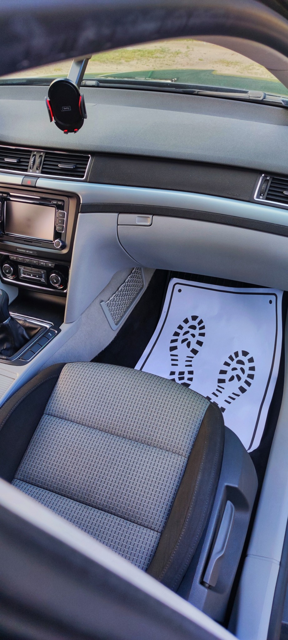 Wnętrze samochodu z szarą tapicerką fotela i papierową matą podłogową z nadrukowanymi śladami butów, widoczne elementy deski rozdzielczej i uchwyt na telefon.