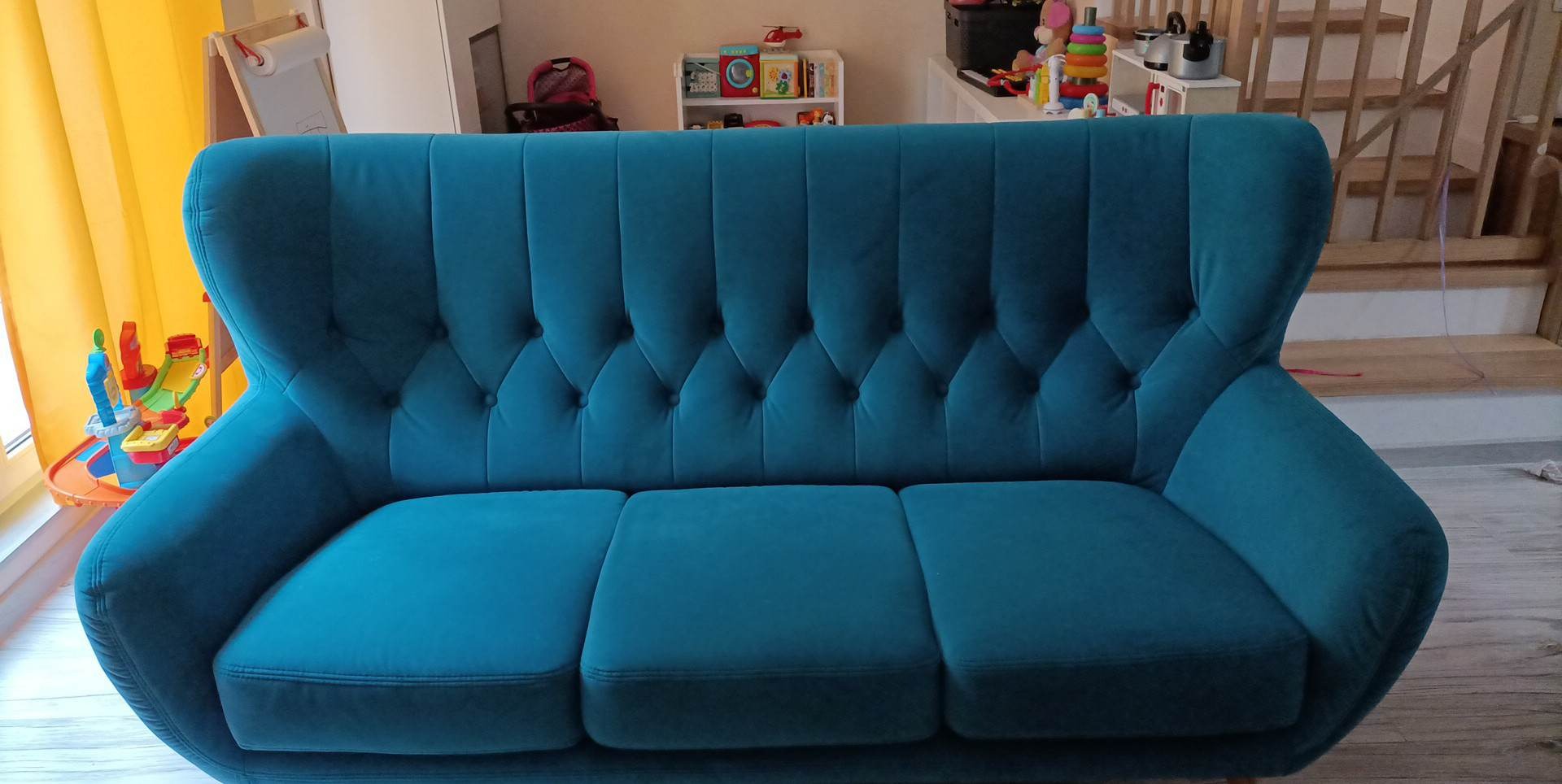 Turkusowa sofa trzyosobowa z pikowanym oparciem w salonie, widoczne zabawki dziecięce w tle.