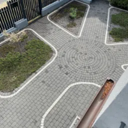 Widok z góry na ułożoną szarą kostkę brukową z białymi obramowaniami, tworzącą wzór rozchodzący się od centralnego okręgu, z elementami zieleni po bokach i widoczną rynną wypełnioną liśćmi.