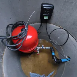 Wnętrze betonowej studni z zamontowanym czerwonym zbiornikiem hydroforowym, czarnym sterownikiem pompy oraz instalacją hydrauliczną z niebieskimi rurami.