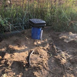 WoDa-mian - Czarna skrzynka sterująca na niebieskiej rurze wystająca z ziemi, otoczona świeżo przekopaną glebą i ogrodzeniem z siatki w tle.