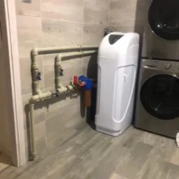 Biały zmiękczacz wody z instalacją hydrauliczną na tle szarych płytek i pralki automatycznej.