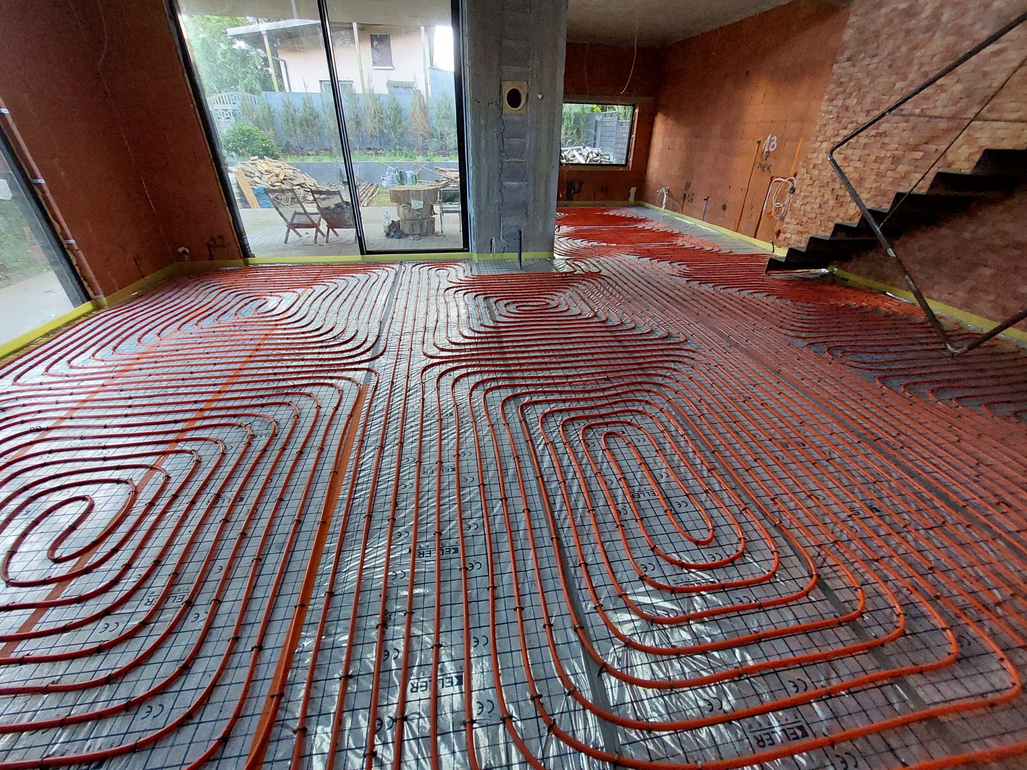 Pomarańczowe rury ogrzewania podłogowego ułożone na folii z siatką, widoczne elementy konstrukcyjne domu w surowym stanie i okno z widokiem na zewnątrz.